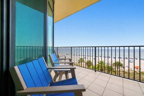 A balcony with two blue and brown lounge chairs, a yellow awning, and a view of the beach with umbrellas.
