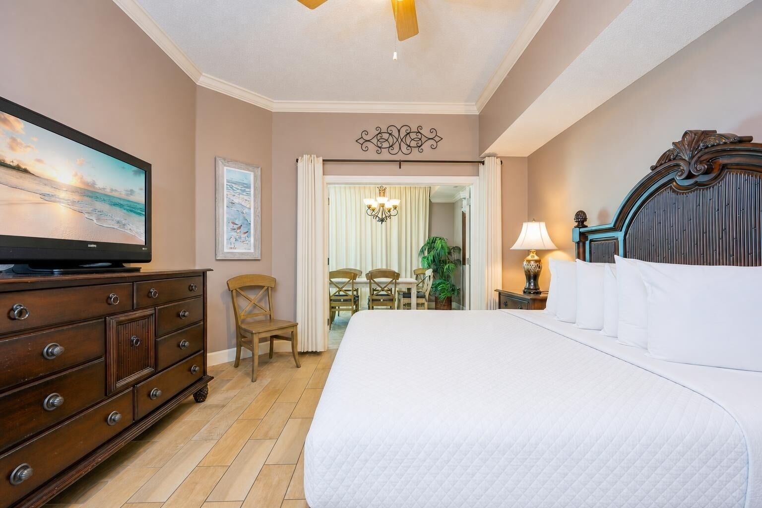 A hotel room with a bed, a television, a wooden dresser, a desk with chairs, and a view of the beach.