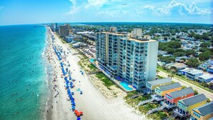 A beachfront hotel with multiple floors, a pool, and a beach with umbrellas. - One Ocean Place 1102 2 Bedrooms, 2 Bathrooms, Oceanfront (Garden City Beach)