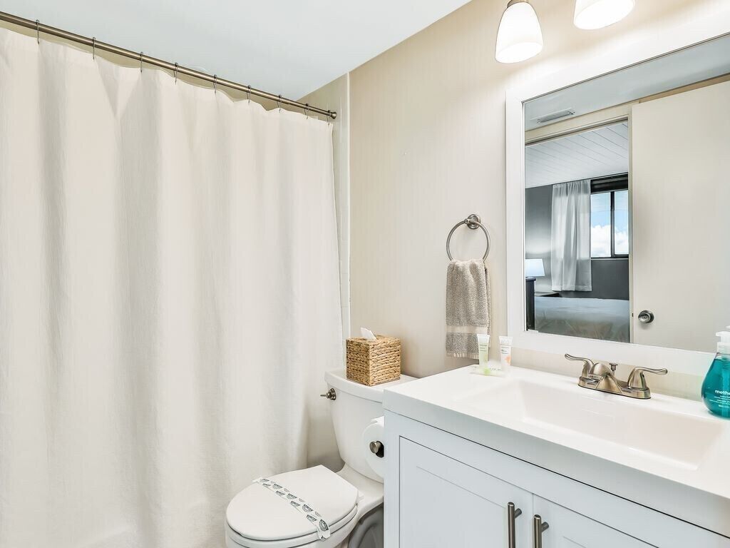 Un baño con inodoro blanco, lavamanos blanco y un espejo encima, y cortina de ducha.