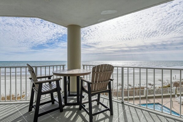 A balcony with a table and two chairs, overlooking a beach with a pool. - One Ocean Place 201 - Spacious three-bedroom, three-bath oceanfront condo (Garden City Beach)