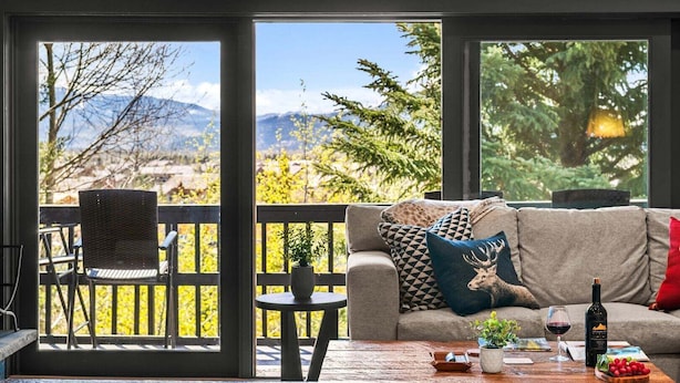 A living room with a sofa, a table with a bottle and glasses, and a view of mountains and trees through large windows. - RMR: Four Seasons I Unit 6 Condo in Teton Village (Teton Village)