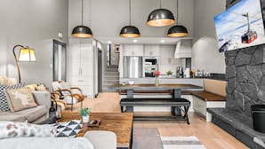 A modern living room with a sofa, coffee table, and a dining area with a bench. There is a kitchen with a stone wall and a dining table. - RMR: Four Seasons I Unit 6 Condo in Teton Village (Teton Village)
