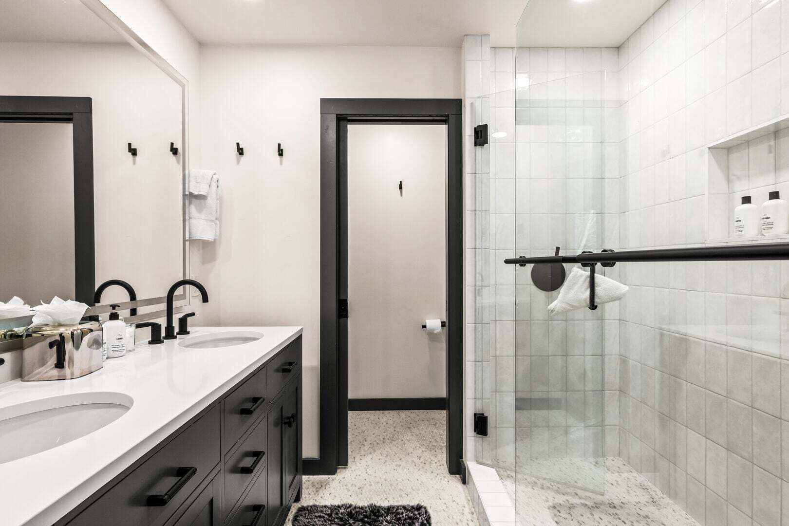 Une salle de bain moderne avec deux lavabos, une douche en verre et une porte sombre donnant sur une autre pièce.