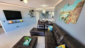 A modern living room with a large flat-screen TV, a leather sofa, and a dining area with a table and chairs.