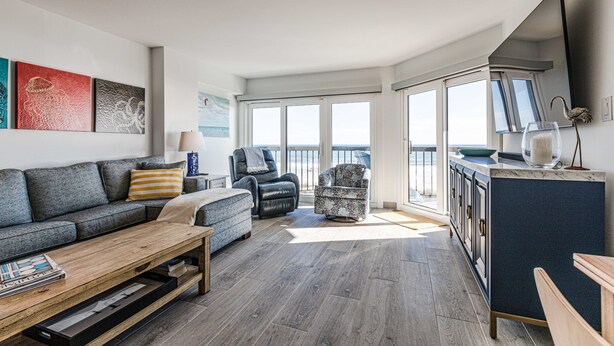 Een moderne woonkamer met een grijze bank, een salontafel en een fauteuil met een patroon. Er is een keuken met een blauwe kast en uitzicht op het balkon.