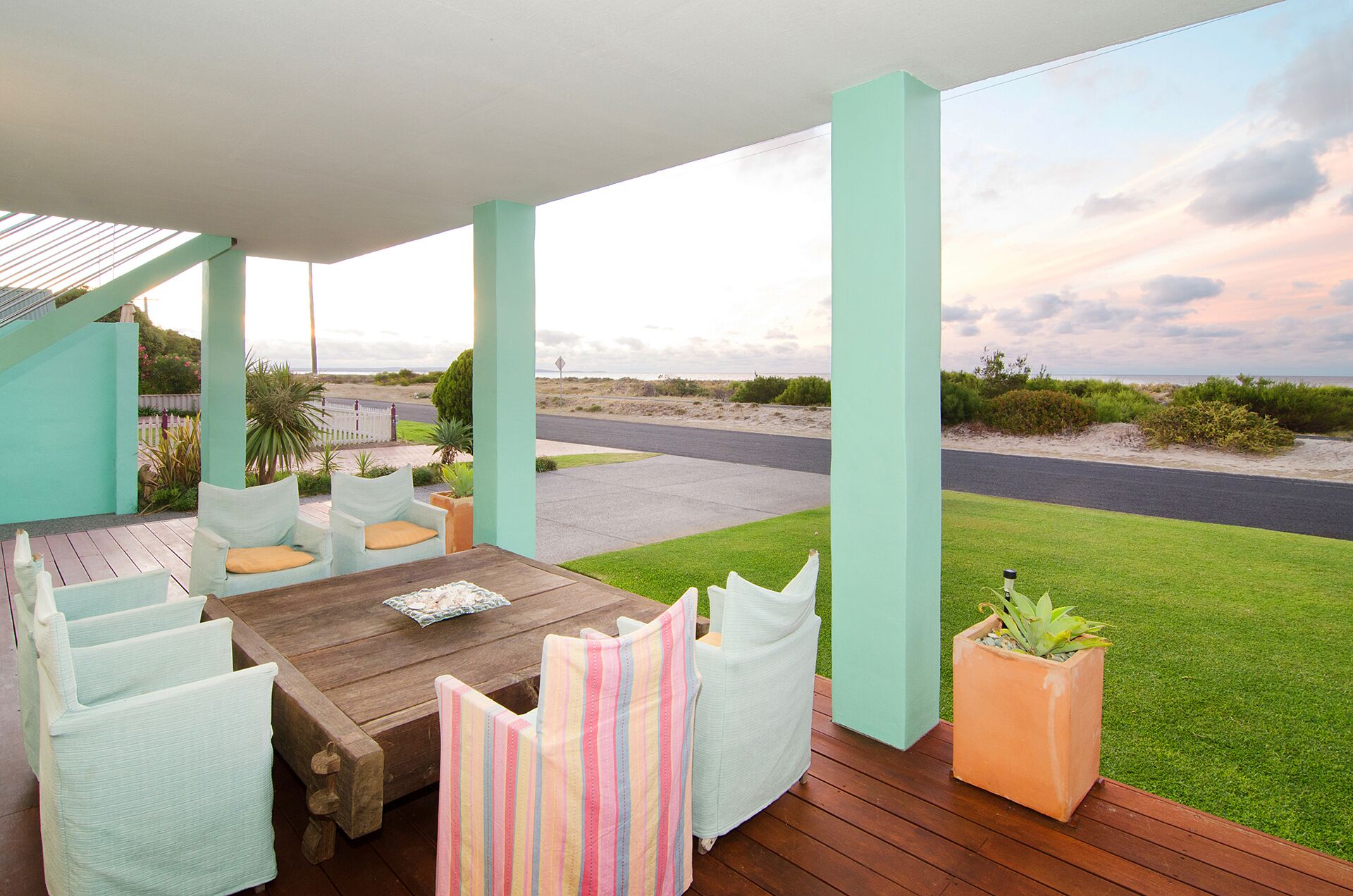 A covered outdoor seating area with a wooden table and cushioned chairs, overlooking a road and grassy landscape.