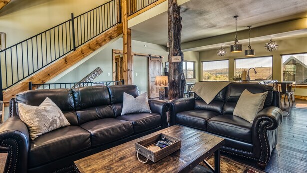 A spacious living area with a black leather sofa, wooden coffee table, and a staircase with wooden railings. - Boat Slip Included, Lake Views, Close to SDC! (Branson)
