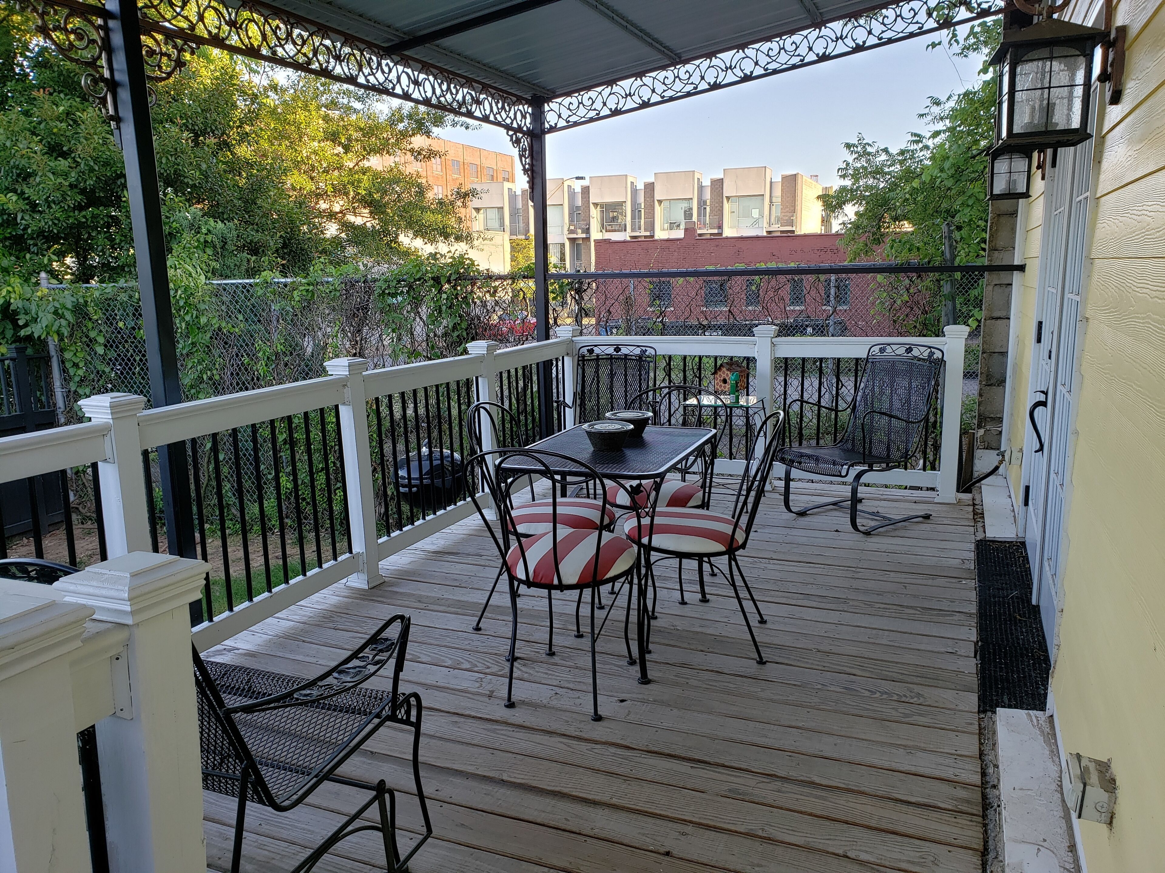 A covered outdoor patio with a table and chairs, surrounded by a white railing and a metal fence.