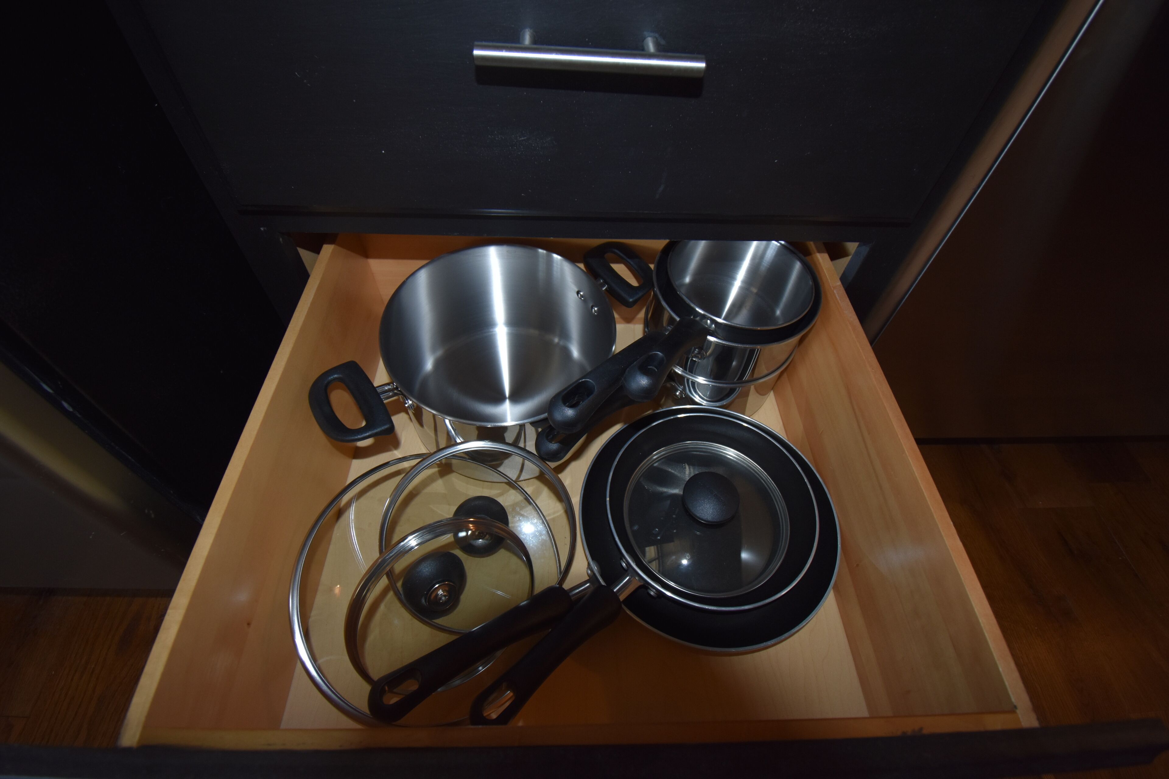 Un cajón de cocina con varios utensilios de cocina, incluyendo ollas, sartenes y una tapa de vidrio.