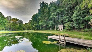 Un étang paisible avec une jetée en bois, entouré d’une végétation luxuriante et d’un bâtiment en pierre.