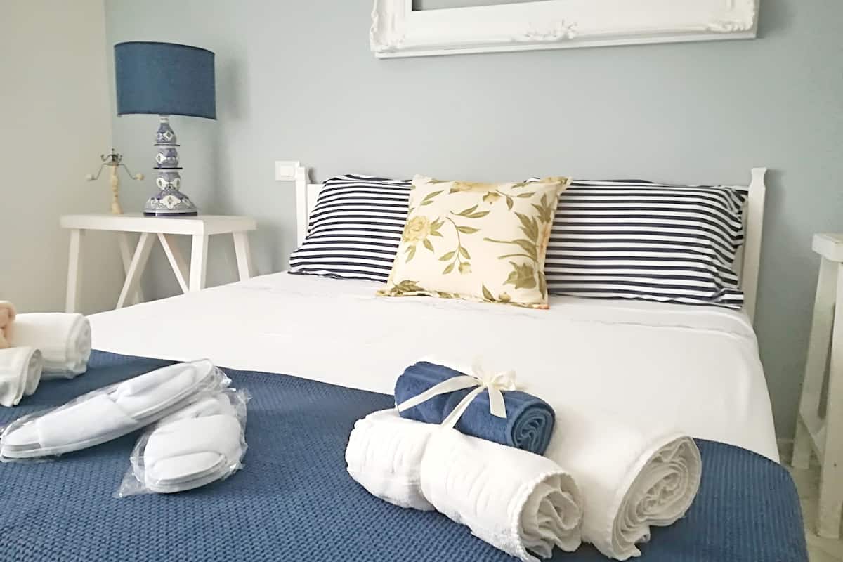 A neatly made bed with striped and floral pillows, a blue bedspread, and rolled towels on a textured blue carpet.