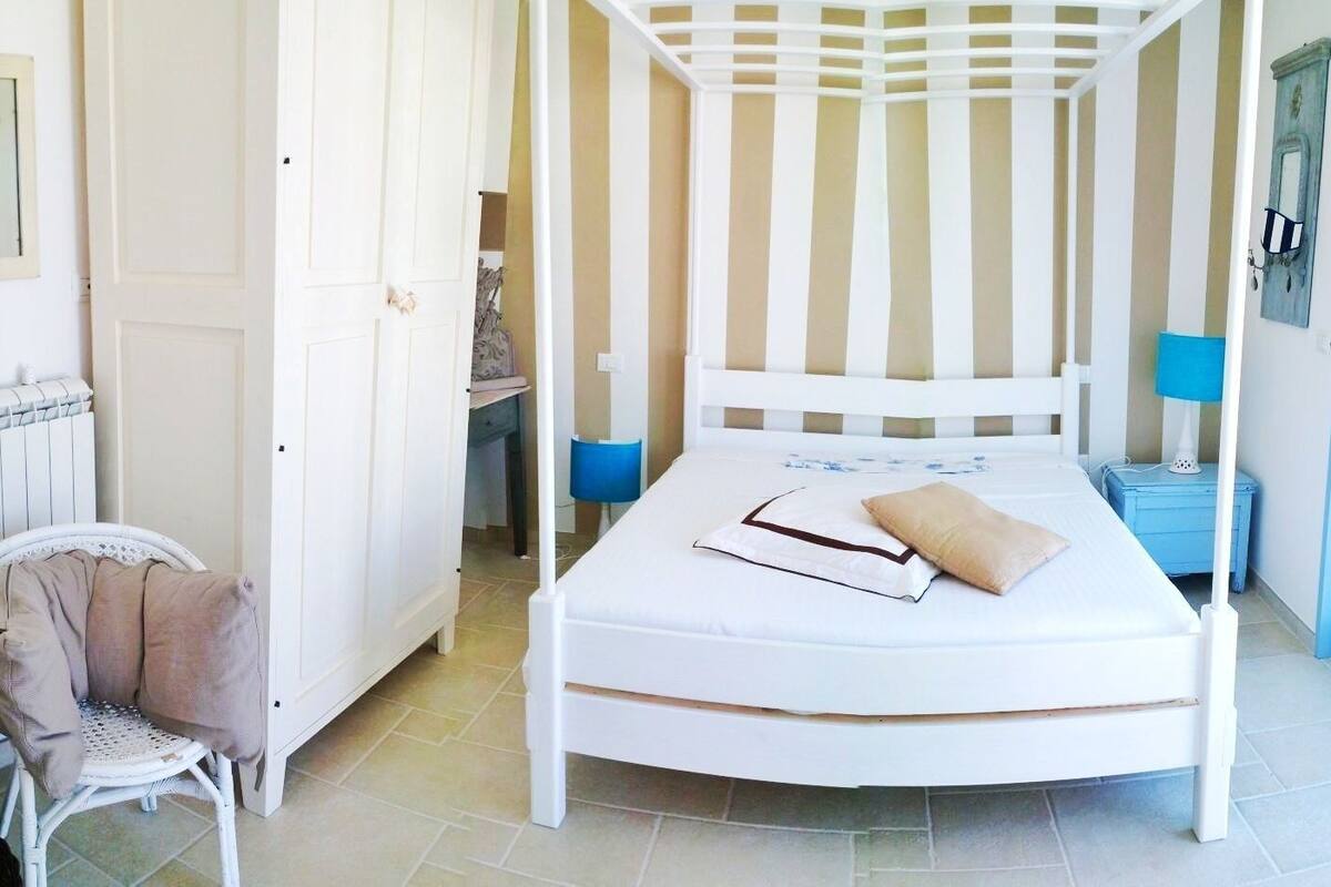 A white four-poster bed with a beige pillow, a chair with a beige cushion, and a small table with a blue lamp.