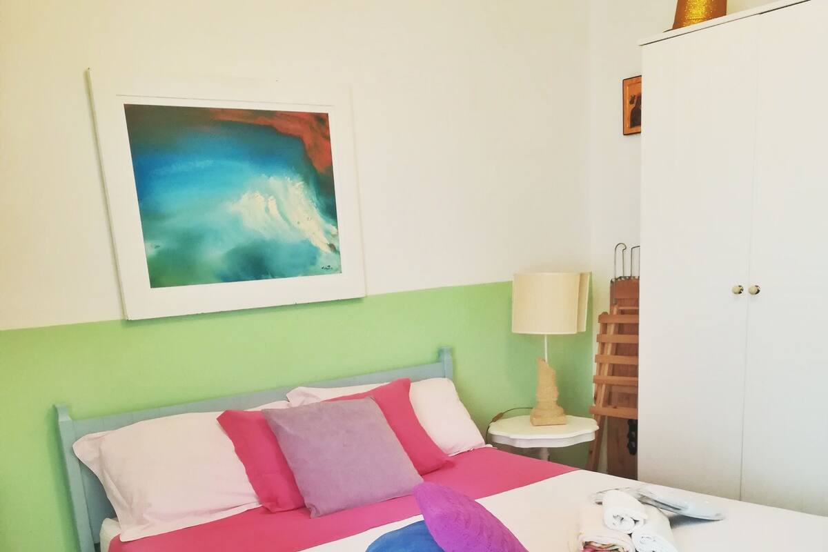 A bed with a blue and pink bedspread, a blue blanket, and a purple pillow. There is a white bedside table with a lamp, a wooden ladder, and a white wardrobe.