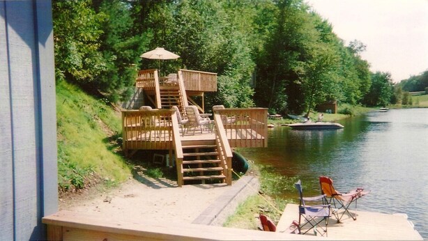 Eine Holzterrasse mit einer Treppe zu einem überdachten Bereich, umgeben von üppigem Grün und einem ruhigen Fluss.