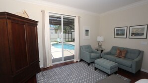 A room with a wooden wardrobe, a grey patterned rug, a grey sofa, a small table with a lamp, and a view of a swimming pool through a glass door. - Lido Key Home w Pool Near Beach & Shops: Rondy Home by RVA (Sarasota)