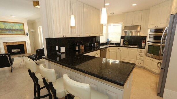 A modern kitchen with white cabinetry, a black granite countertop, and stainless steel appliances. There are three bar stools and a dining table in the adjacent area. - Lido Key Home w Pool Near Beach & Shops: Rondy Home by RVA (Sarasota)