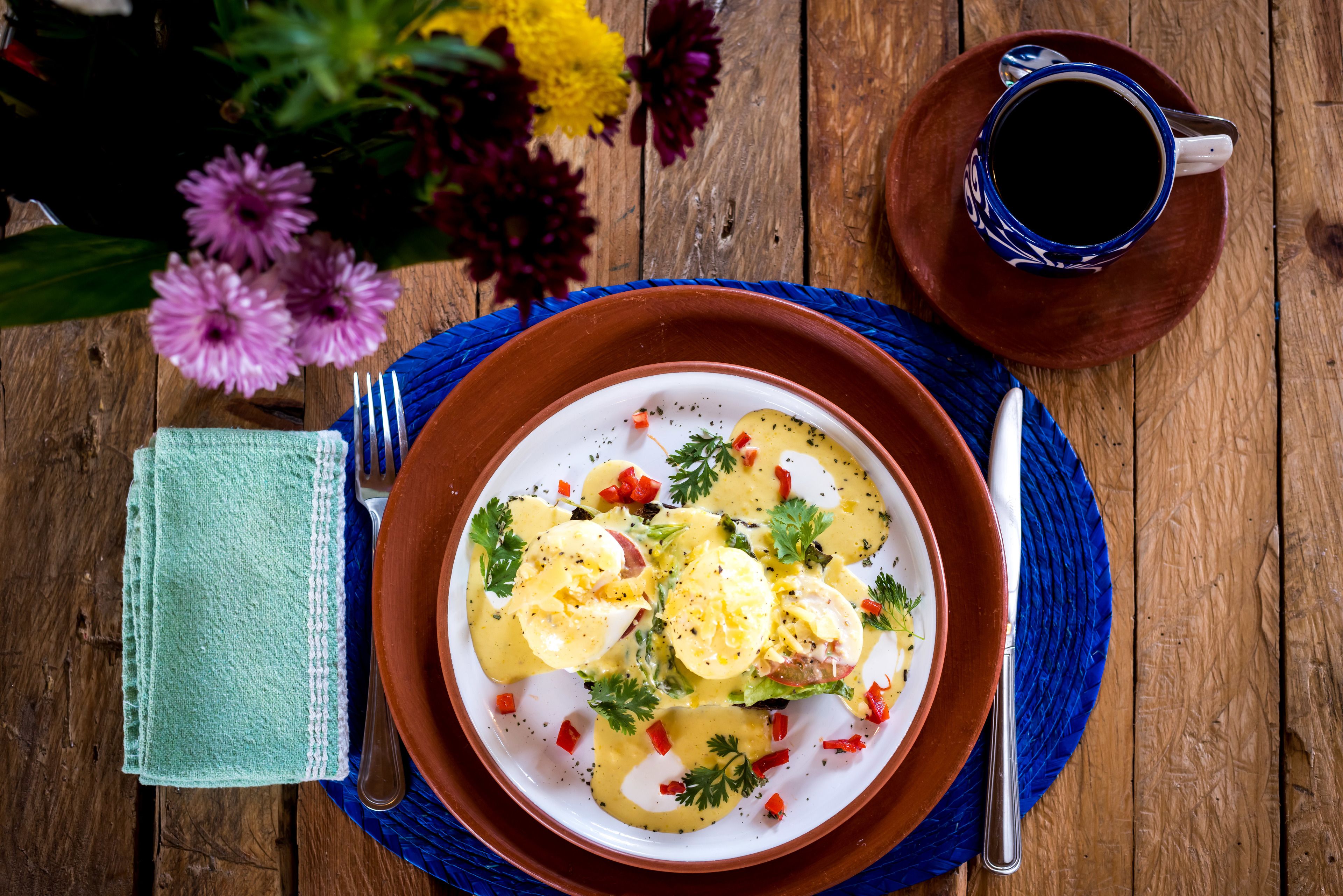 A plate of food with eggs, greens, and red peppers, a cup of coffee, and a vase of flowers on a wooden table.