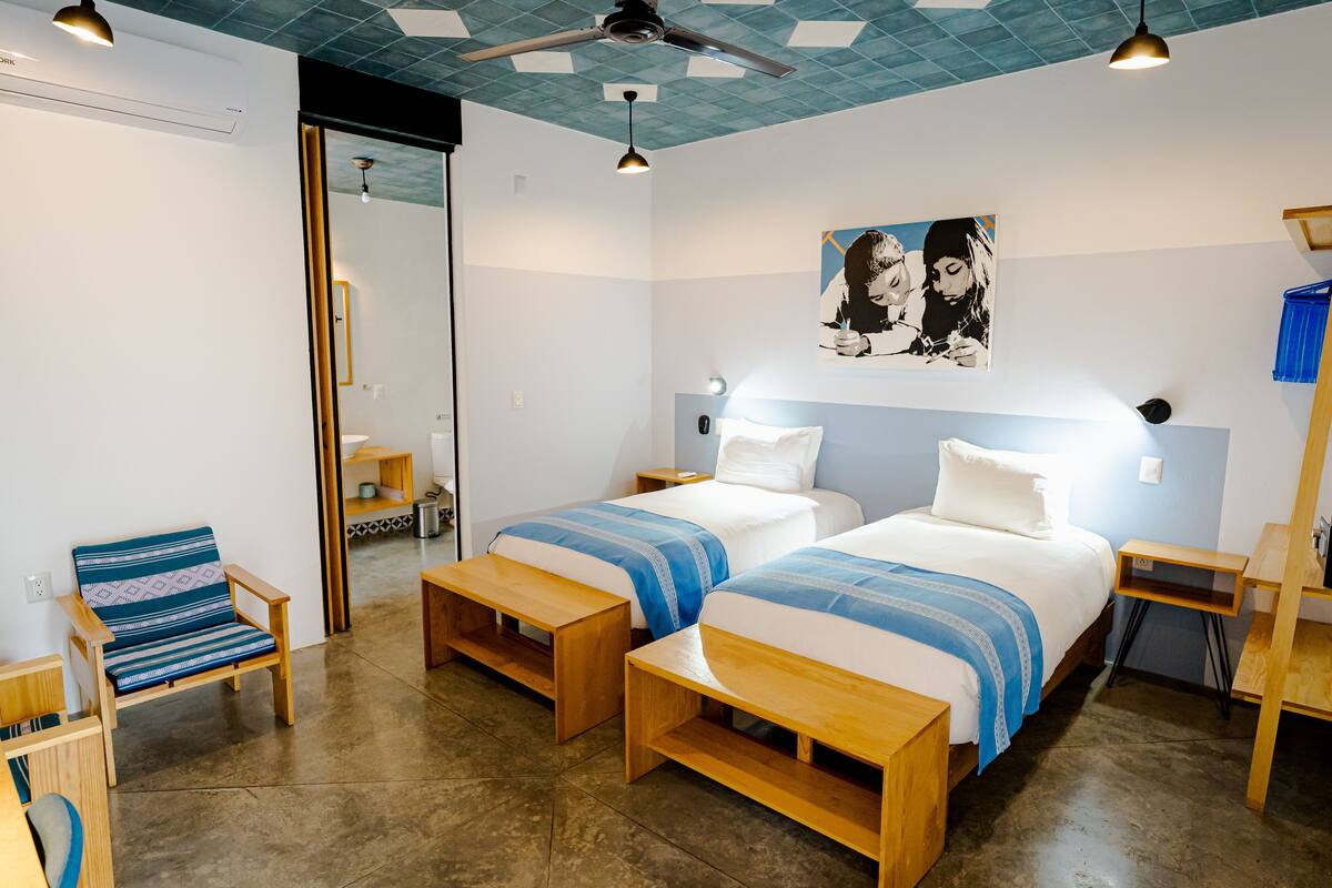 A hotel room with two beds, a chair, a bench, a ceiling fan, and a painting on the wall.