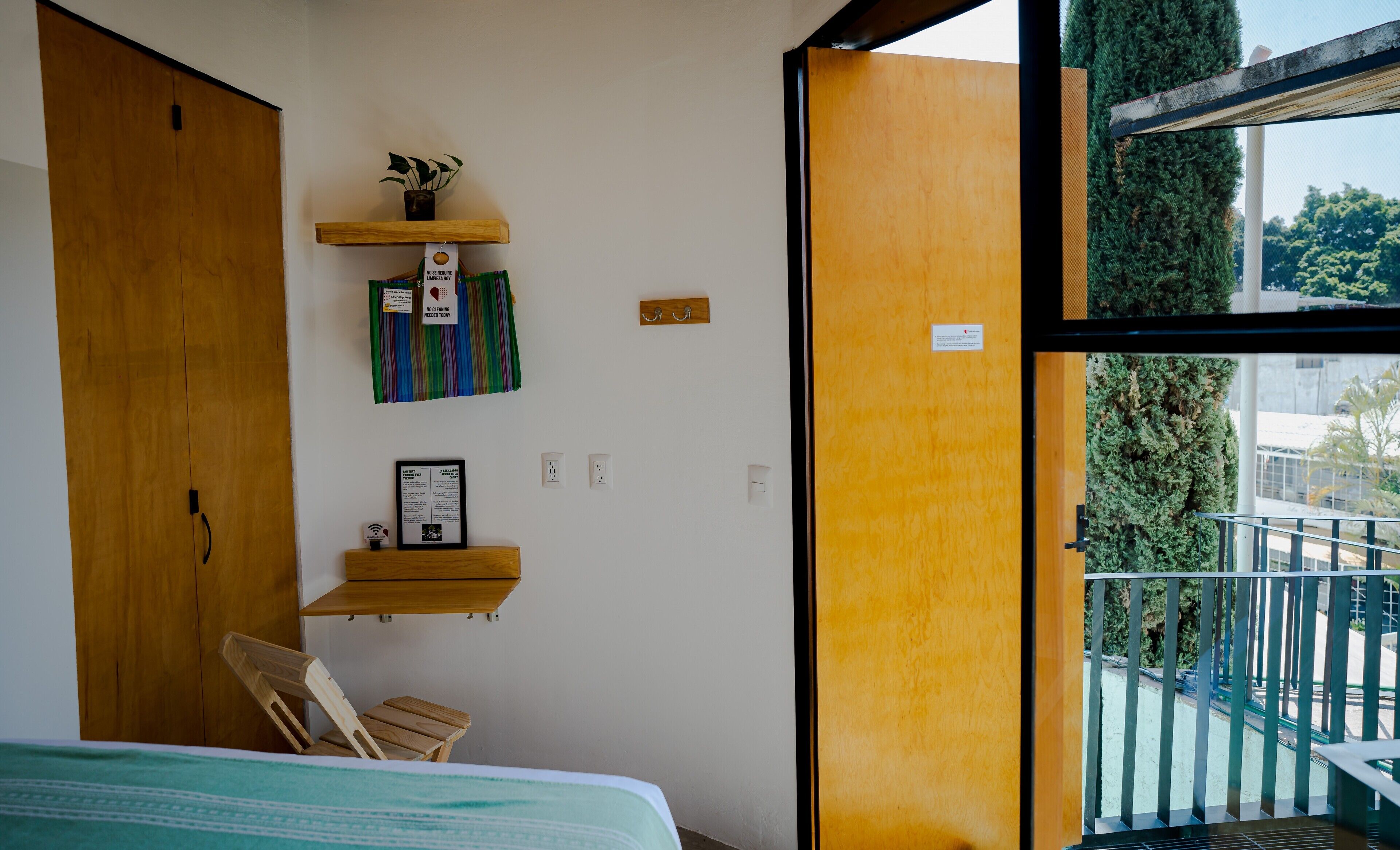 Un dormitorio con cama, armario de madera, una pequeña mesa de madera, una silla, ventana con vistas a zonas verdes y balcón con barandilla.