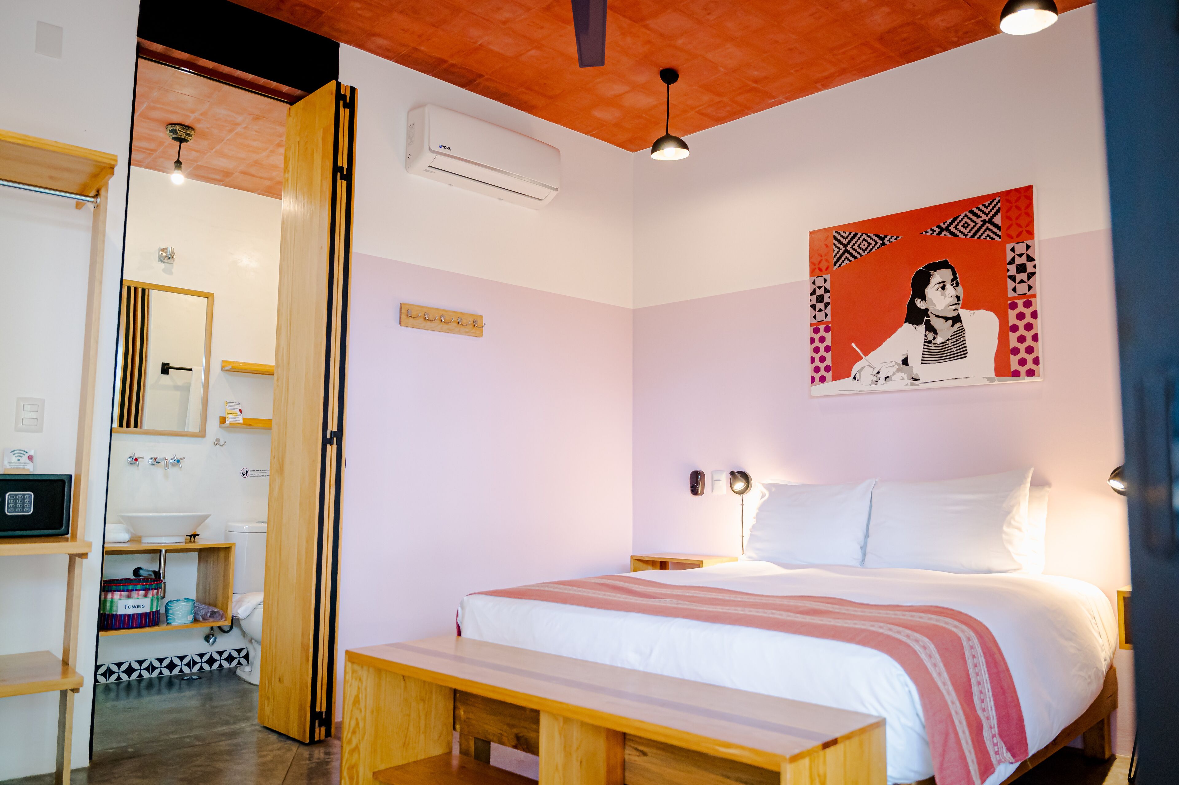 A bedroom with a bed, bedside table, and a bench. There is a bathroom with a toilet, sink, and mirror. A ceiling fan and an air conditioning unit are visible.