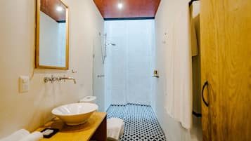 Un baño con tocador de madera, lavamanos blanco, espejo y zona de ducha.