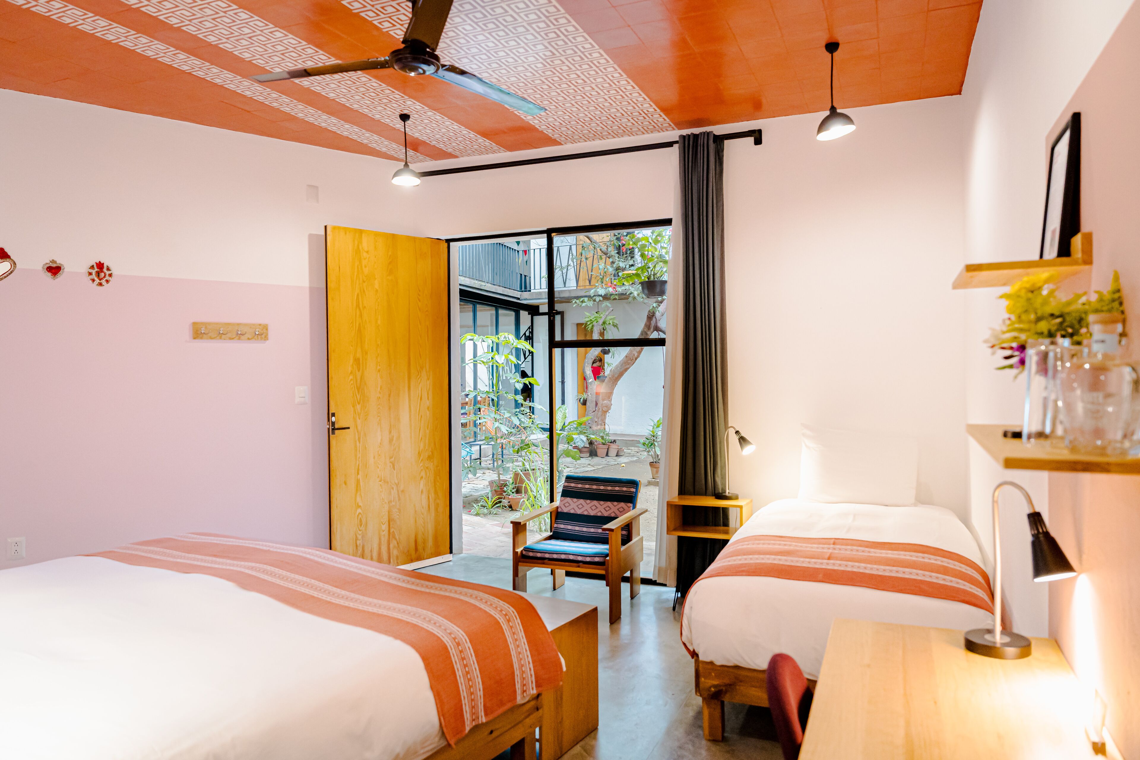 A hotel room with two beds, a wooden ceiling, a ceiling fan, a window with a view of the outside, a small table with a lamp, and a shelf with decorative items.