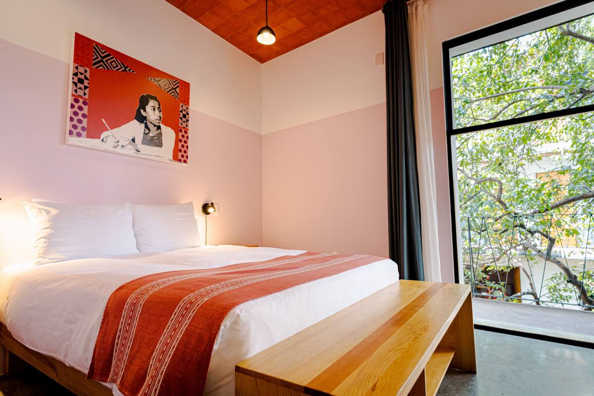 A bedroom with a large bed, a wooden bench, a painting on the wall, and a window with a view of trees.