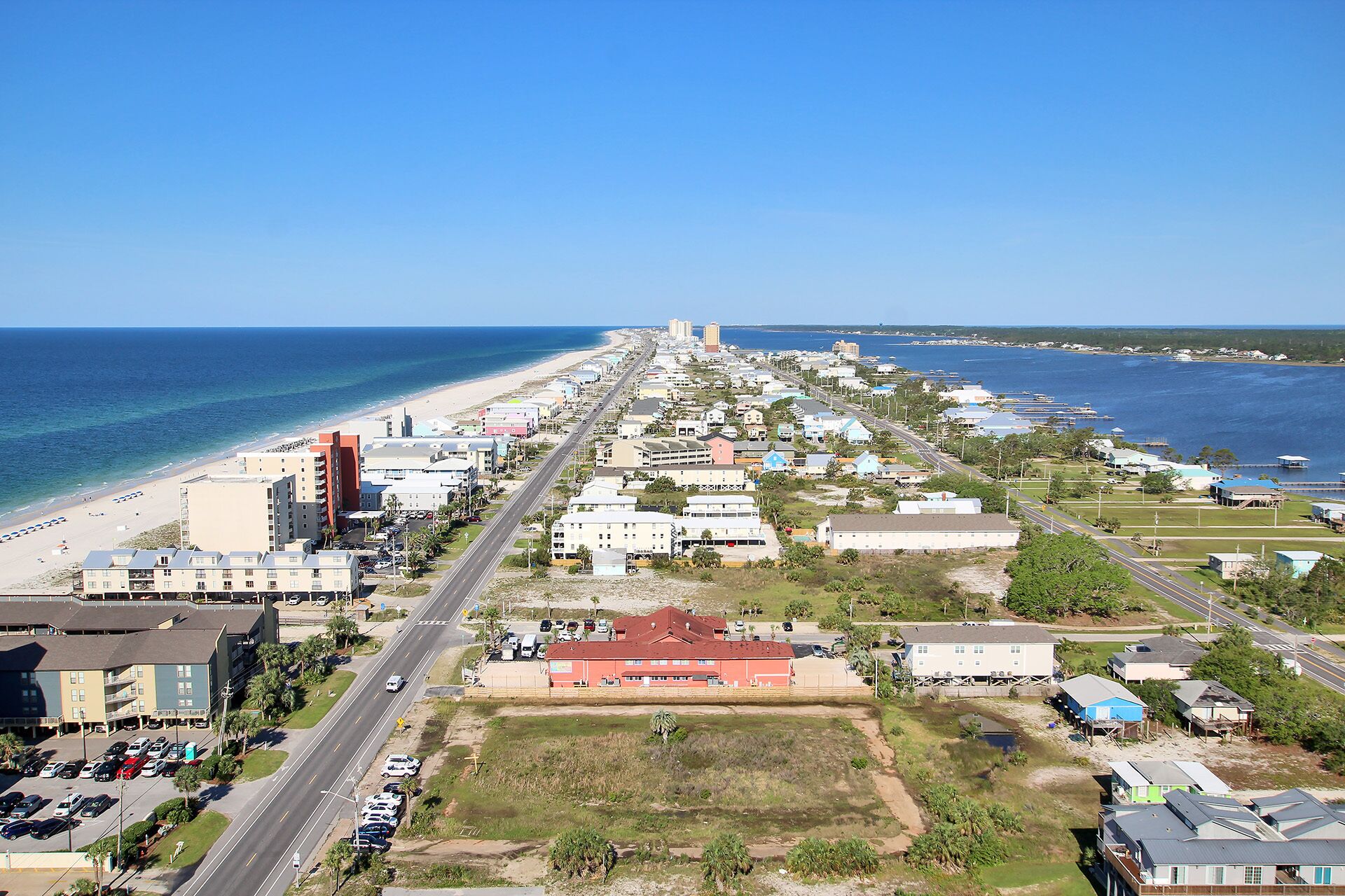 An aerial view of a coastal town with a beach, a main road, and various buildings including a prominent red building in the center.
