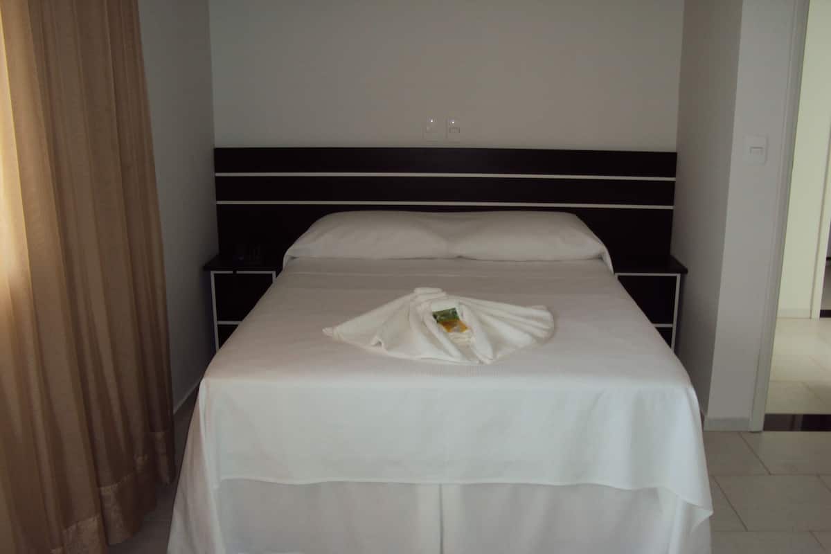 Una cama bien tendida con colcha y almohadas blancas, una mesita de noche con lámpara, y una cortina entreabierta.