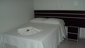 A single bed with a white bedspread and pillows, a bedside table with a black object on it, and a white wall with a power outlet. - Laf Hotel (Registro)