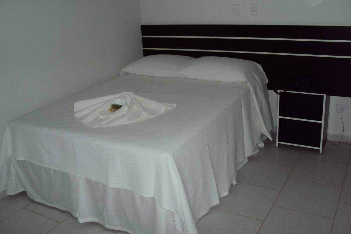 Una cama individual con edredón y almohadas blancos, una mesita de noche con un objeto negro encima y una pared blanca con un enchufe.