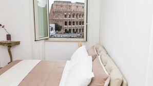 Ein Schlafzimmer mit Bett, einem Fenster mit Blick auf ein römisches Amphitheater aus der Antike, einem kleinen Tisch mit Vase und einer Lampe.