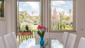 Ein Essbereich mit einem Glastisch, weißen Stühlen und Blick auf einen Garten mit Bäumen und Blumen.