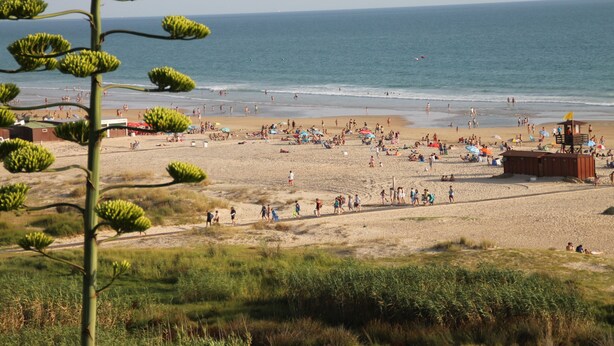 Une plage avec des gens et des parasols, une tour de surveillance des sauveteurs, et un ciel dégagé.