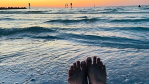 Pies en el agua al atardecer, con una vista distante de un muelle y una persona en el agua.