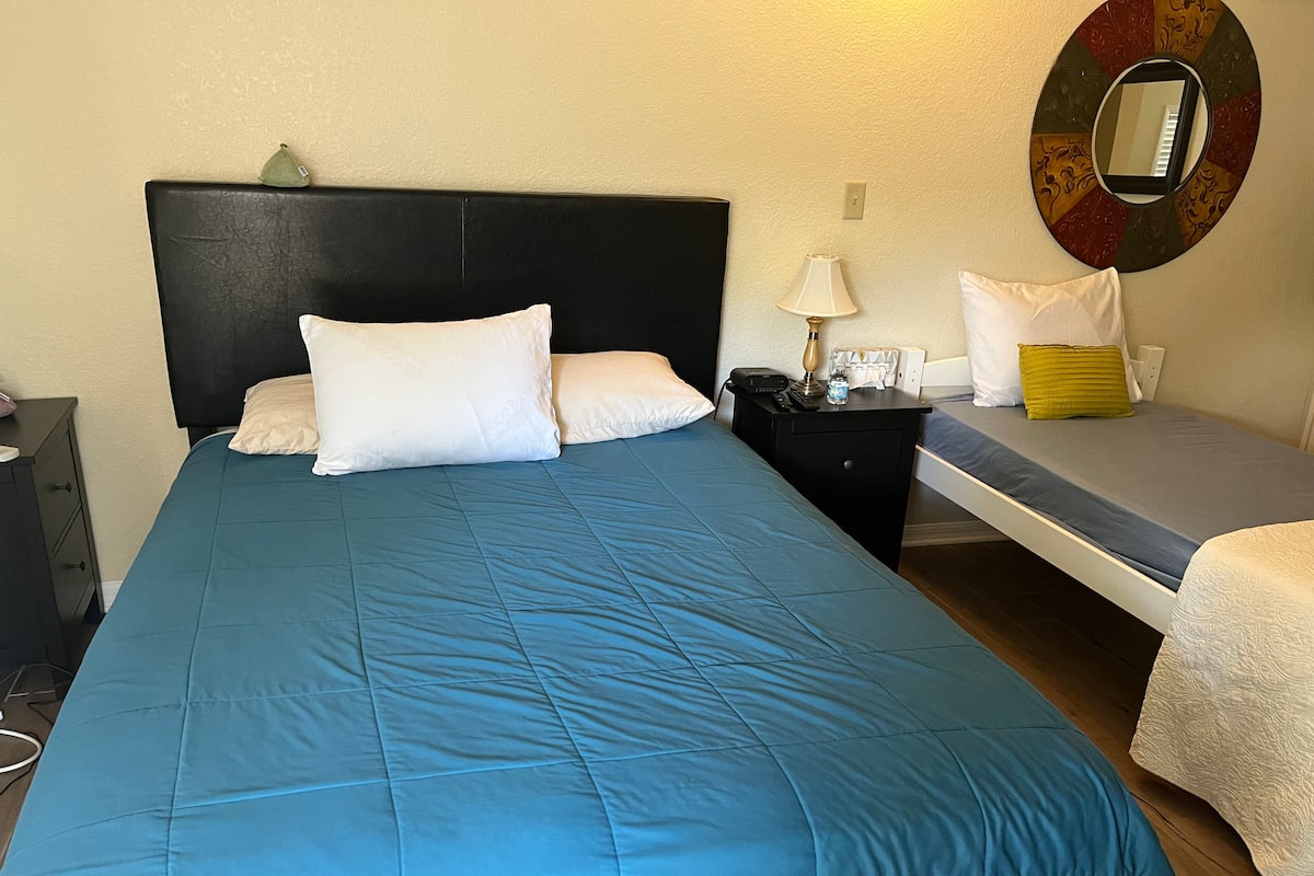 Una cama bien hecha con una colcha azul y almohadas blancas, una mesita de noche con una lámpara, una planta pequeña y un espejo fijado a la pared.