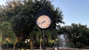 Un parque con un poste de reloj, bancos, una fuente y una bicicleta.