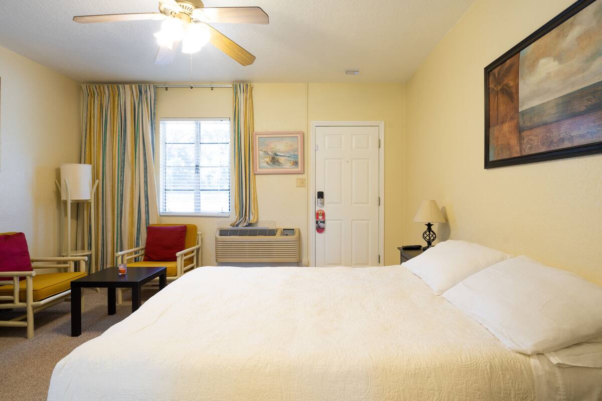Habitación de hotel con una cama grande, un ventilador de techo, una ventana con cortinas, un cuadro en la pared y una mesita de noche con una lámpara.