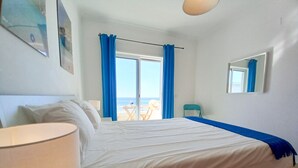 Une chambre d’hôtel avec un lit, une chaise, une table, une fenêtre avec des rideaux et une vue sur la plage.