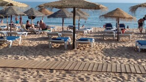 Une plage avec des chaises longues et des parasols en chaume, une promenade en bois, et des gens qui profitent de la plage.
