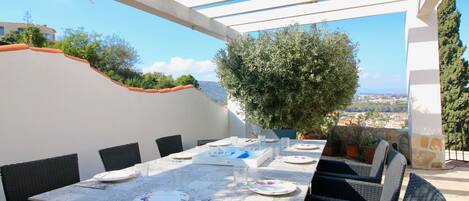 An outdoor dining area with a table set for four, surrounded by wicker chairs, under a white pergola with a view of the city and hills.