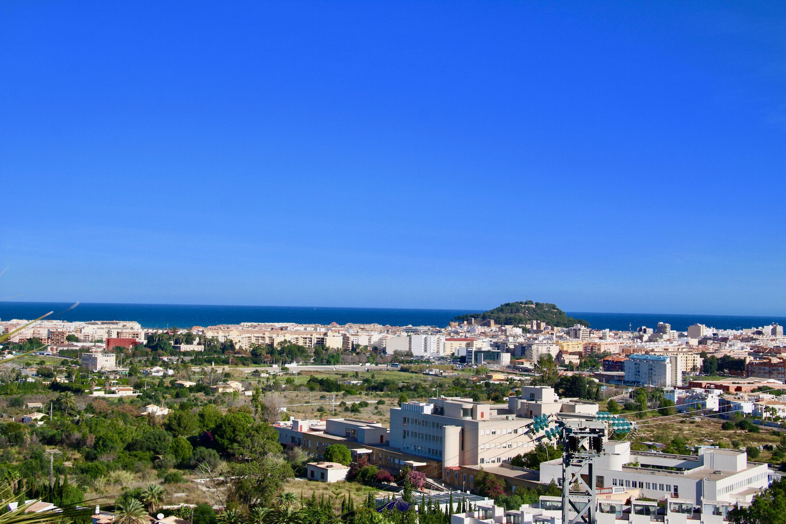 A coastal cityscape with a mix of residential and commercial buildings, a prominent hill, and a clear blue sky.