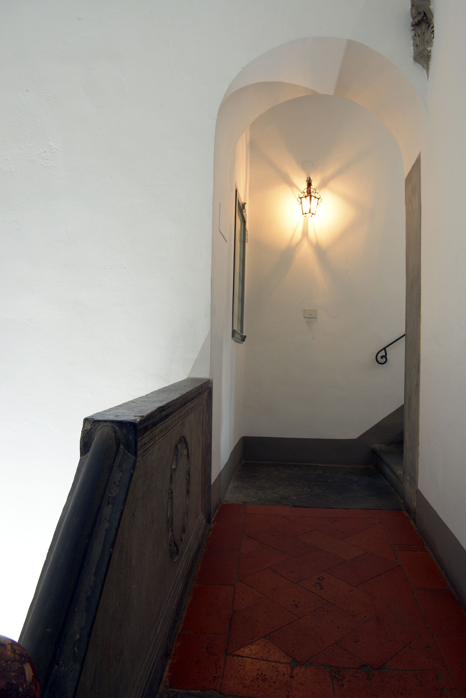 Un escalier étroit avec un sol recouvert de carreaux rouges, des murs blancs et un luminaire fixé au mur.