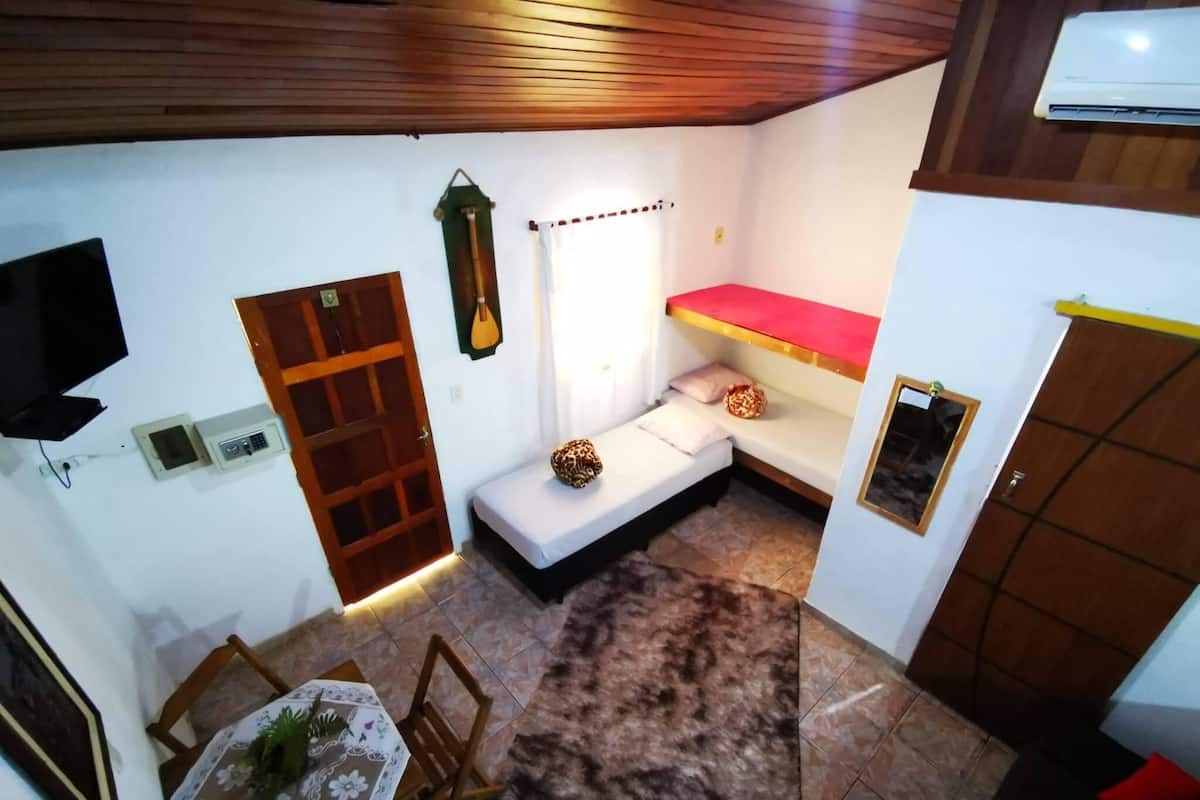 A compact room with a wooden ceiling, a small table with a floral tablecloth, a bench with cushions, a wall-mounted TV, and a door with a security panel.