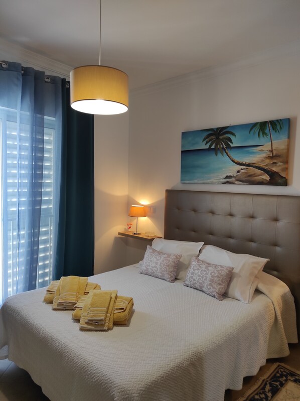 Une chambre d’hôtel avec un lit, un tableau représentant une scène de plage, une lampe de chevet et une fenêtre avec des rideaux.