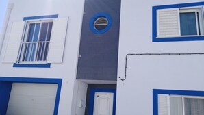 Un edificio con fachada blanca y azul, que incluye una puerta de garaje, una puerta con teclado numérico y una ventana con contraventanas blancas.
