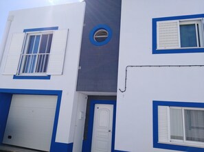 A building with a white and blue facade, featuring a garage door, a door with a keypad, and a window with a white shutter.