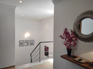 Un escalier avec une rampe, un miroir fixé au mur et une étagère ornée d’un vase de fleurs.