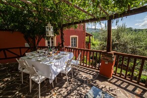Uma área de refeições ao ar livre com uma mesa posta para uma refeição, cercada por uma pérgola de madeira e com vista para um prédio vermelho e vegetação.